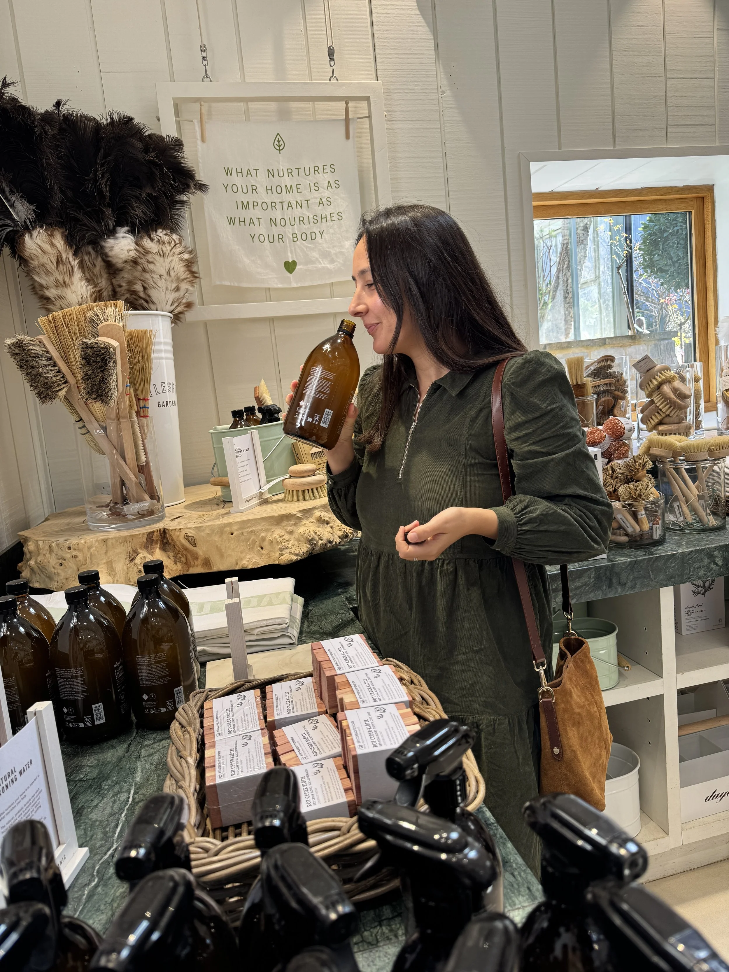 A woman shopping in a store, holding a brown bottle and surrounded by natural products like brushes, soaps, and skincare items, with a decorative sign on the wall.