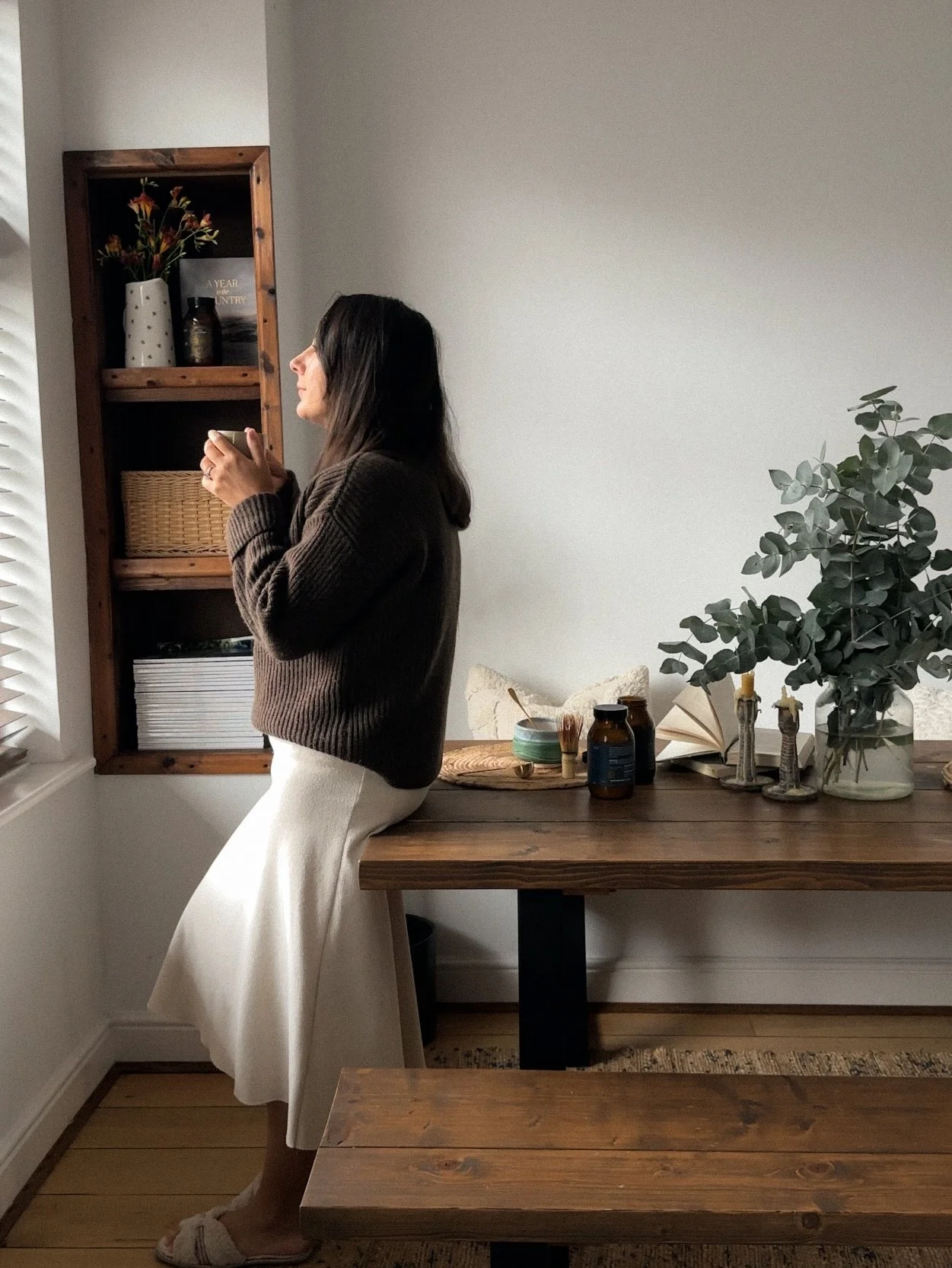 A woman with dark hair, wearing a brown sweater and white skirt, sits on a wooden table holding a mug, in a cozy interior with a wooden shelf, plants, and books.