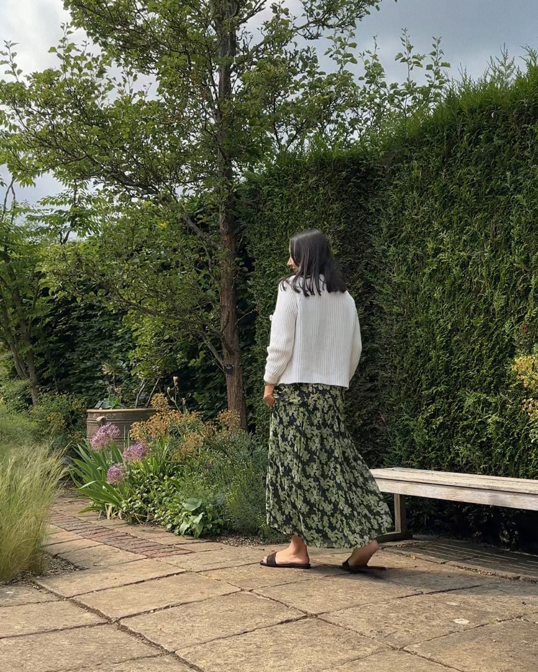 A woman with long dark hair in glasses, wearing a white sweater, a long green floral skirt, and sandals, stands on a garden path near a lush hedge and a tree, with plants and flowers nearby.