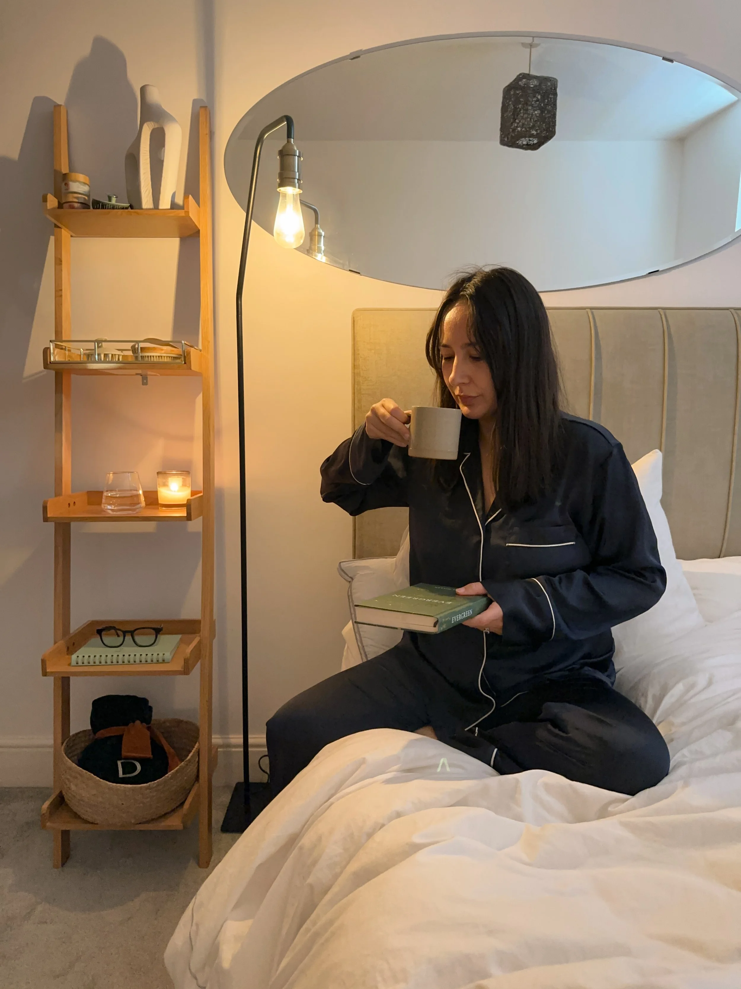 A woman in pajamas sitting on a bed, drinking coffee and holding a book.