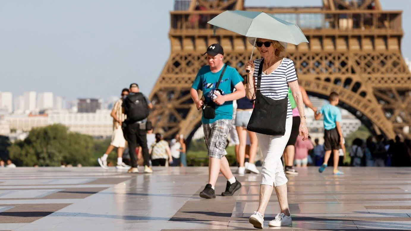 Tourists walking near the Eiffel Tower in Paris during a summer heatwave, some using umbrellas for shade.