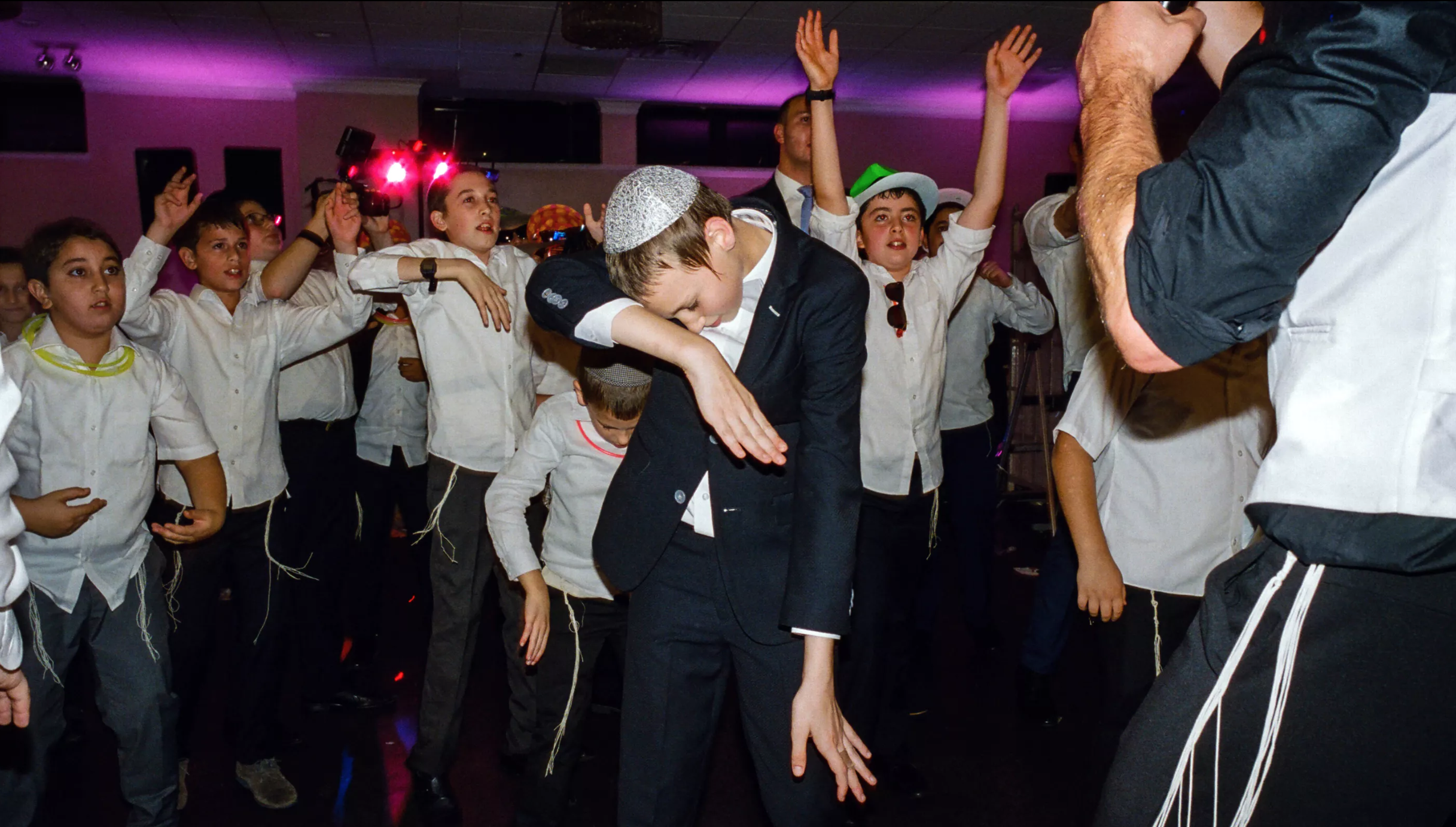 Children dancing and celebrating at a party, with some wearing kippahs and festive attire in a room with purple and pink lighting.