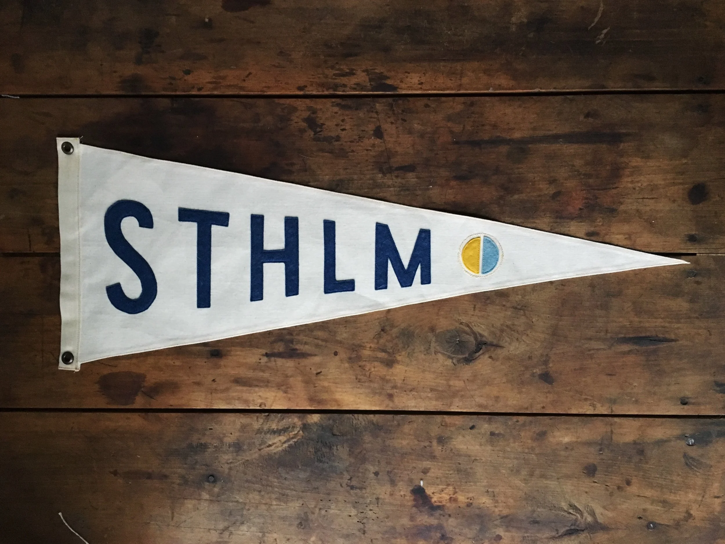 A triangular fabric sign displaying the letters 'STHLM' in navy blue with a small round emblem featuring yellow, blue, and white colors on a wooden surface.
