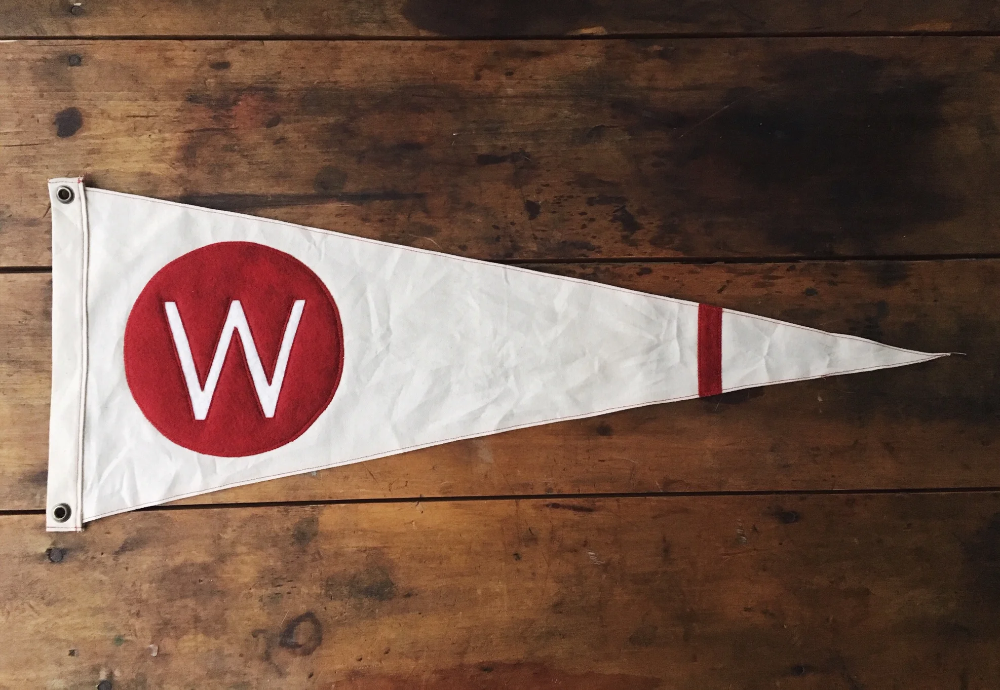 White triangular pennant flag with a large red circle containing a white letter "W" and a small red marking near the tip, placed on a wooden surface.