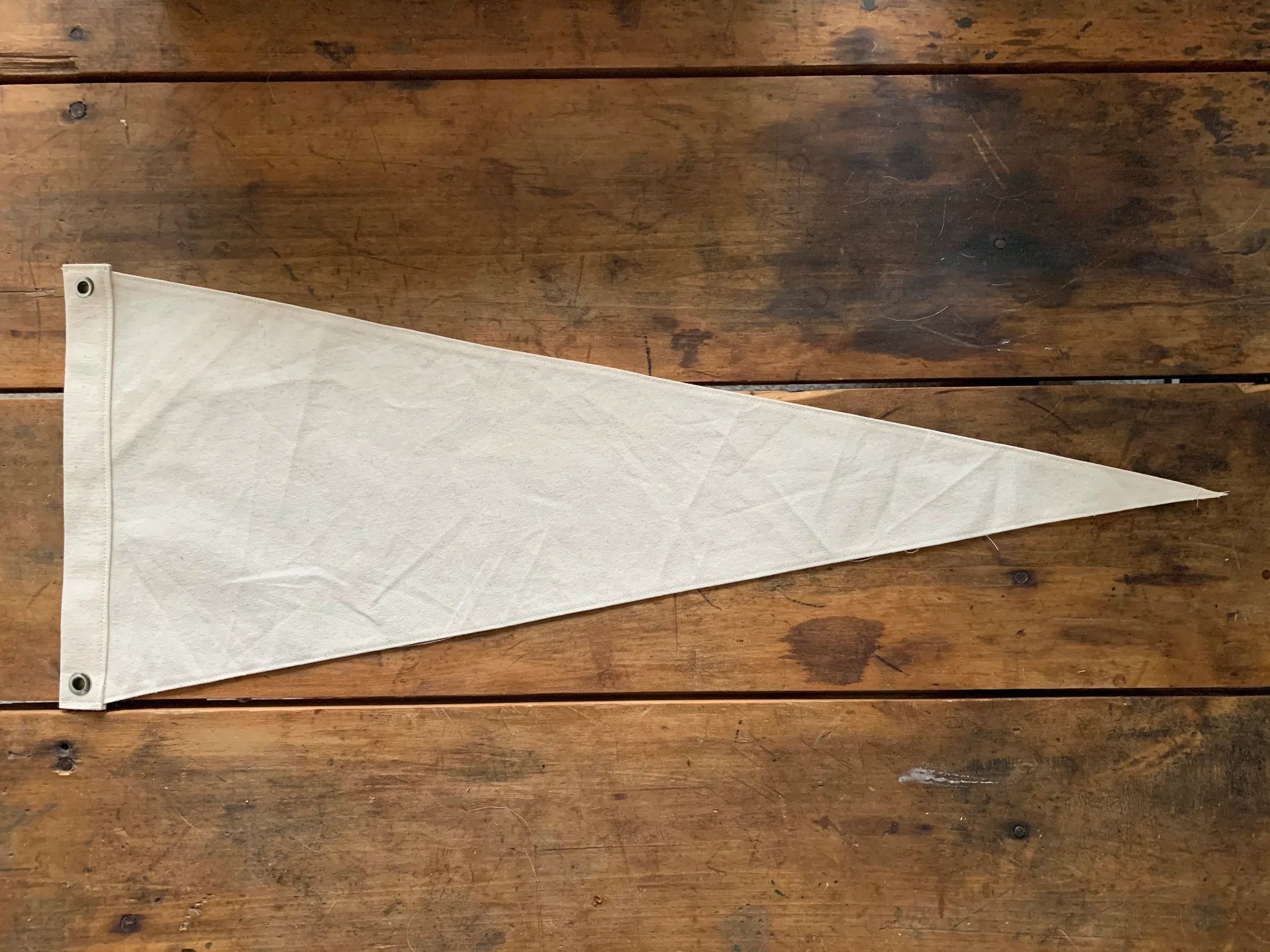 A blank beige pennant flag with metal grommets at the top corners, lying on a wooden surface.