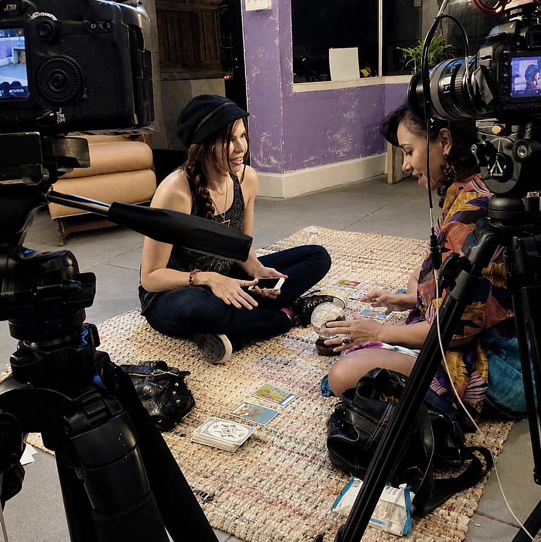 Two women sitting on a rug playing tarot cards, with cameras filming them in an indoor space with purple walls and a window in the background.