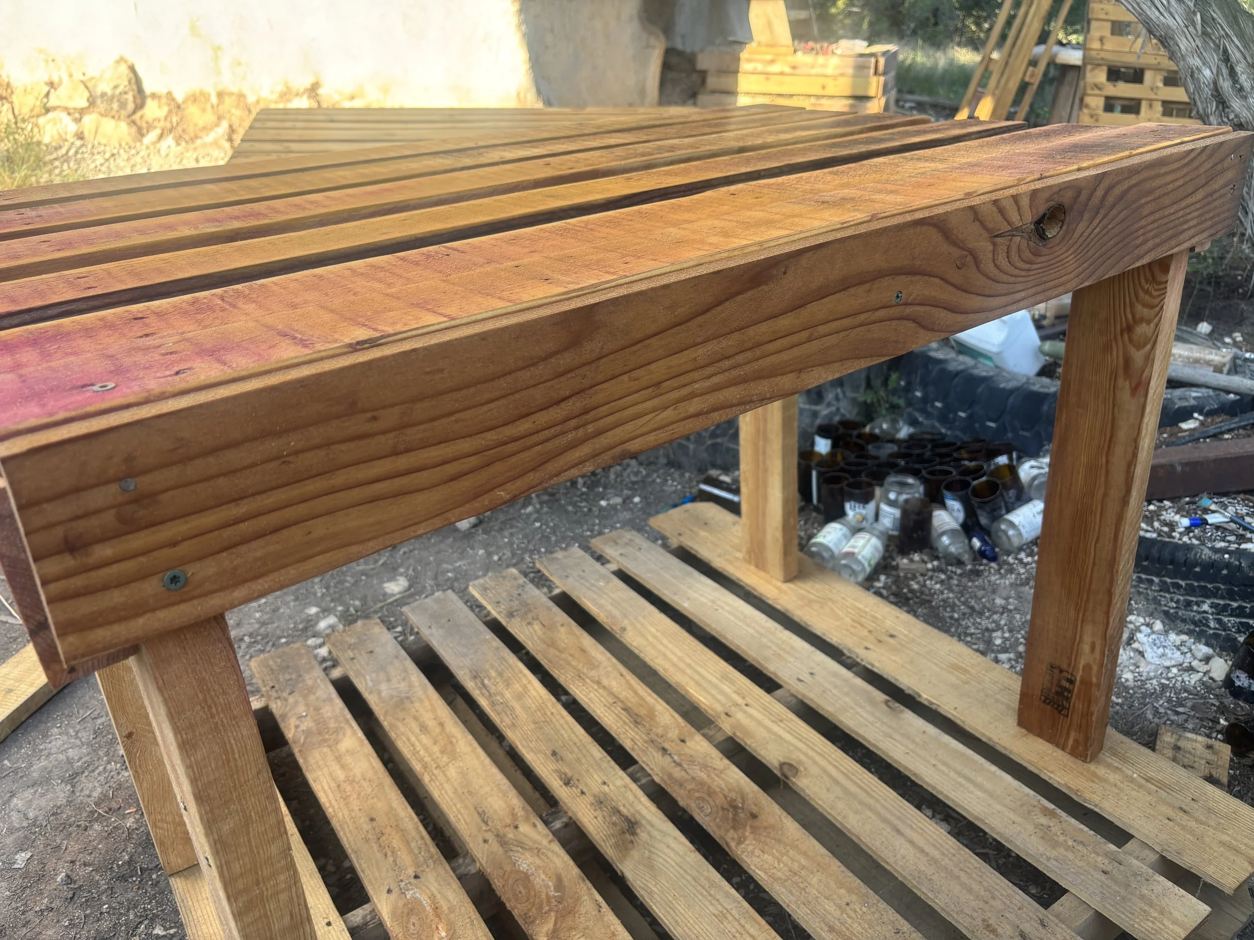 Wooden bench with slats, outside in a construction or workshop area with tools and supplies underneath and around it.