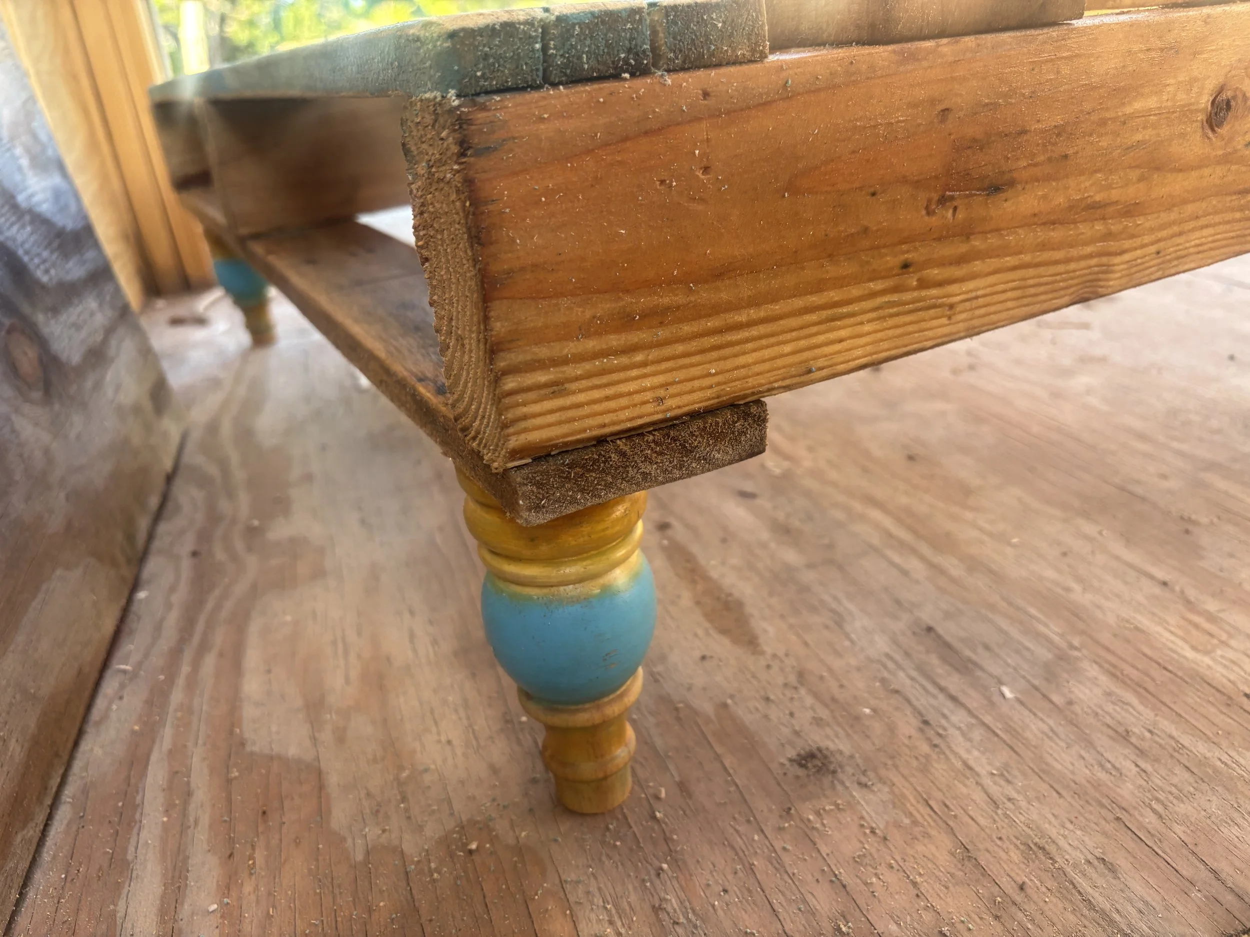 Close-up of a wooden table leg with a turned spindle design, painted in bright colors including yellow and blue, next to a wooden floor and a piece of furniture.