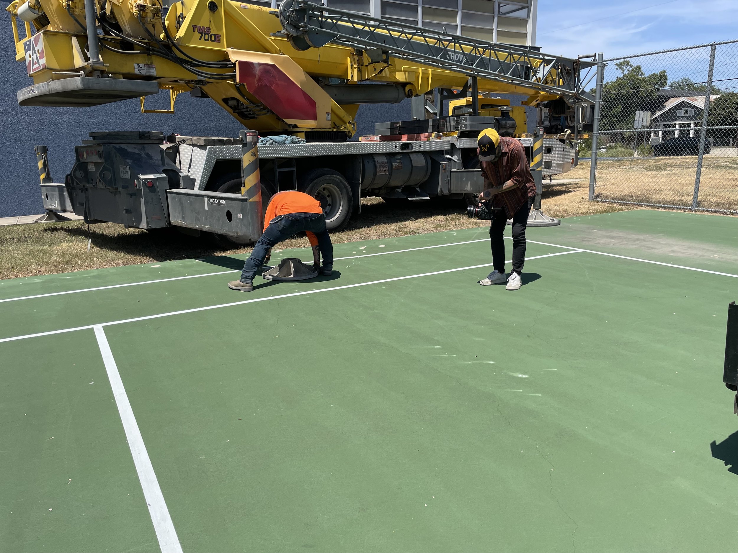 Two crew members working on a sports court with a large yellow construction crane parked on the side. One person is in an orange shirt bending down, while the other is standing with a camera near a fence.