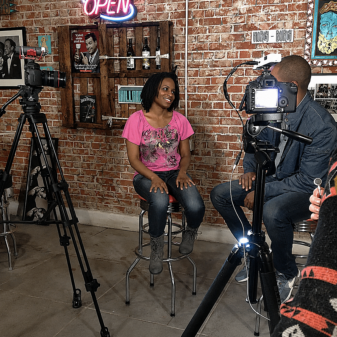 A woman with short, curly black hair wearing a pink shirt with a graphic design, sitting on a stool during an interview in a cafe with exposed brick walls. Two cameras are filming her, and a man is speaking to her during the interview.