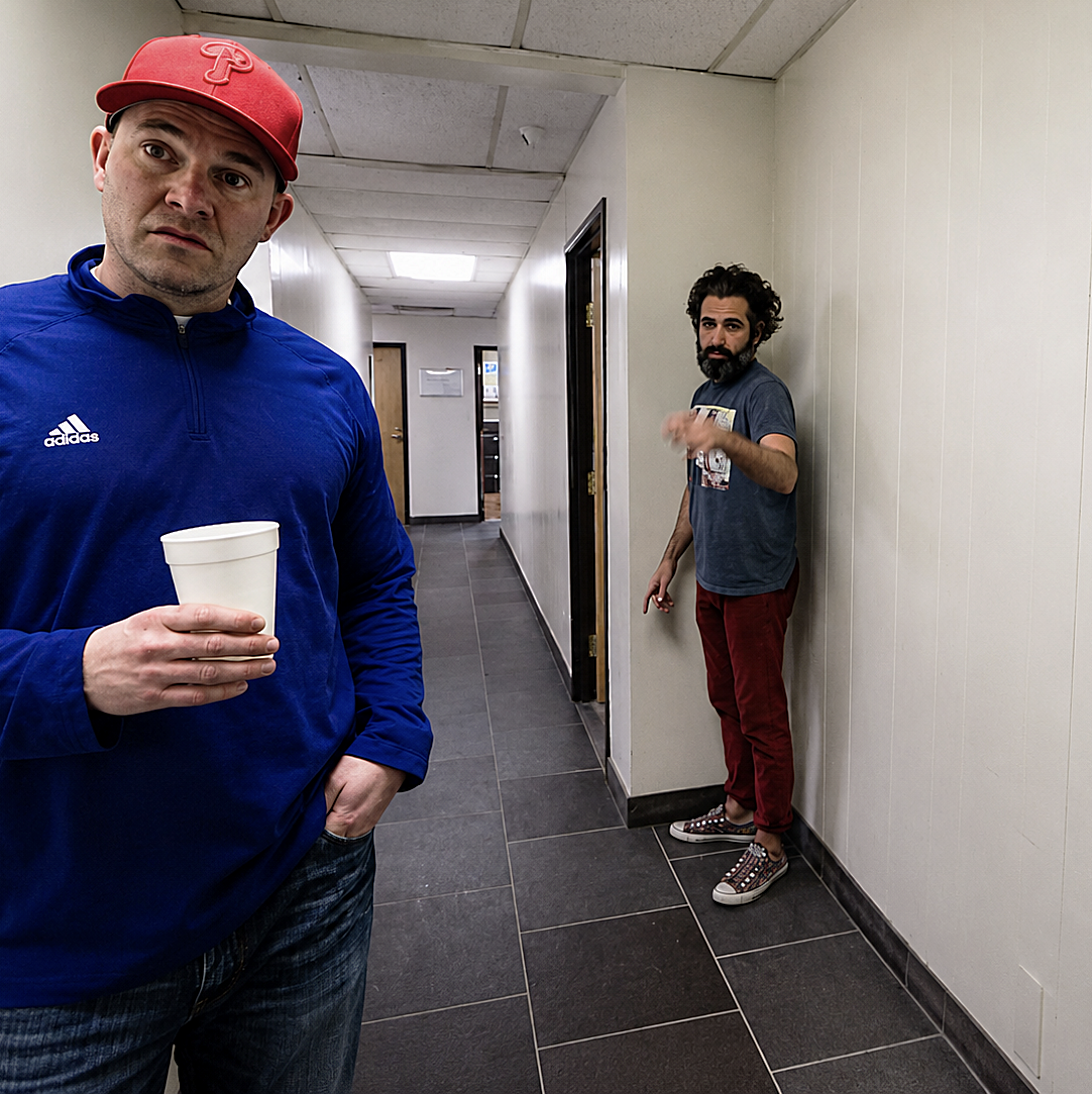 Two men standing in a hallway, one in foreground wearing a red cap and blue jacket holding a white cup, the other in background wearing a gray t-shirt and red pants leaning against the wall, looking towards the camera.