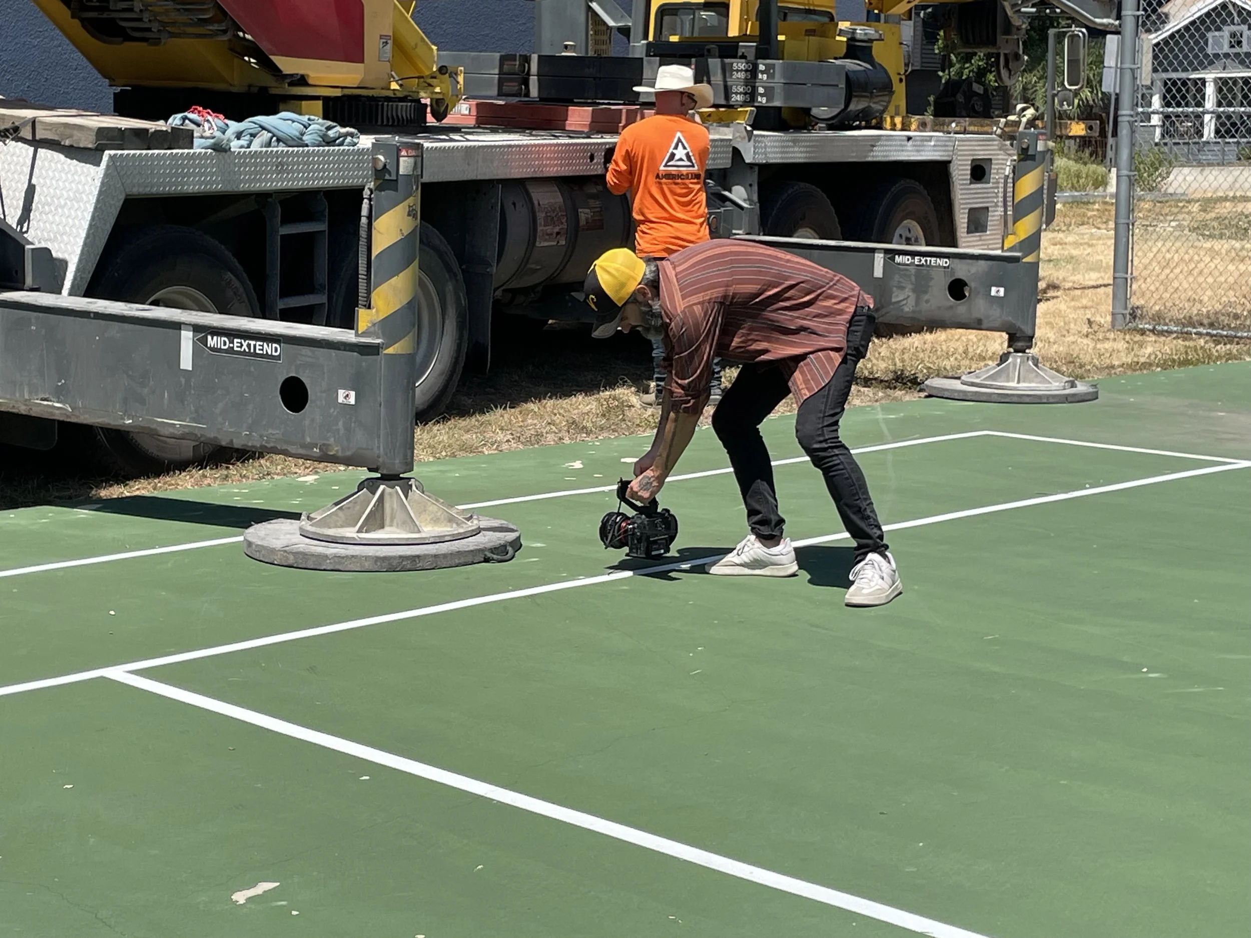 Two workers on a green tennis court are setting up equipment near a large tow truck. One worker is wearing an orange shirt and white cowboy hat, the other is dressed in a plaid shirt and yellow cap. The tow truck's flatbed is loaded with machinery and tools, with a chain and some ropes visible. There is a chain-link fence and trees in the background.