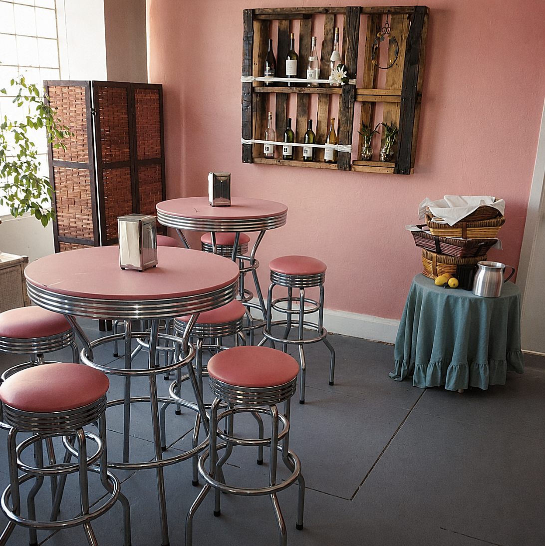 A cozy retro-style dining area with pink tables and matching bar stools featuring chrome accents, a pink wall with wooden shelves displaying wine bottles, and a small round table covered with a teal tablecloth holding wicker baskets, lemons, and a me