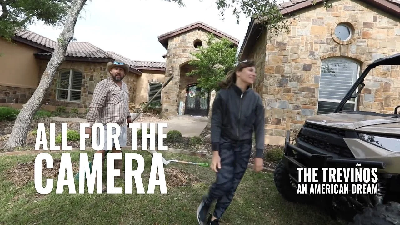 A man and a woman working in the yard of a large stone house, with the woman passing by the man holding a rake, and a large vehicle parked nearby.