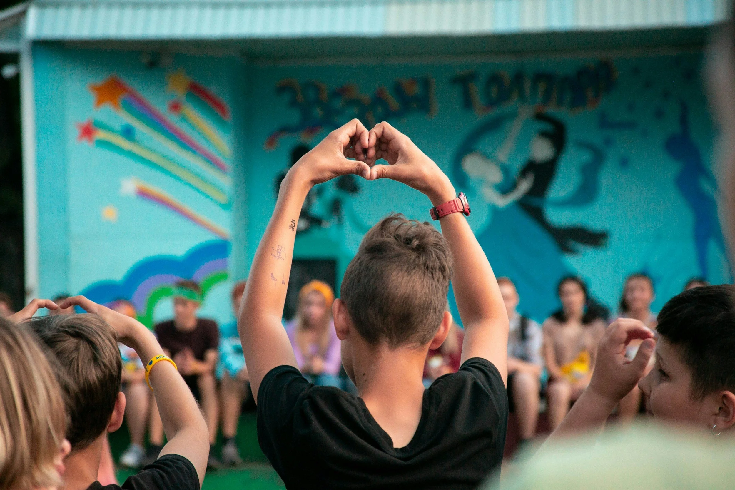 Children in a classroom raising their hands to participate, with colorful wall art in the background.