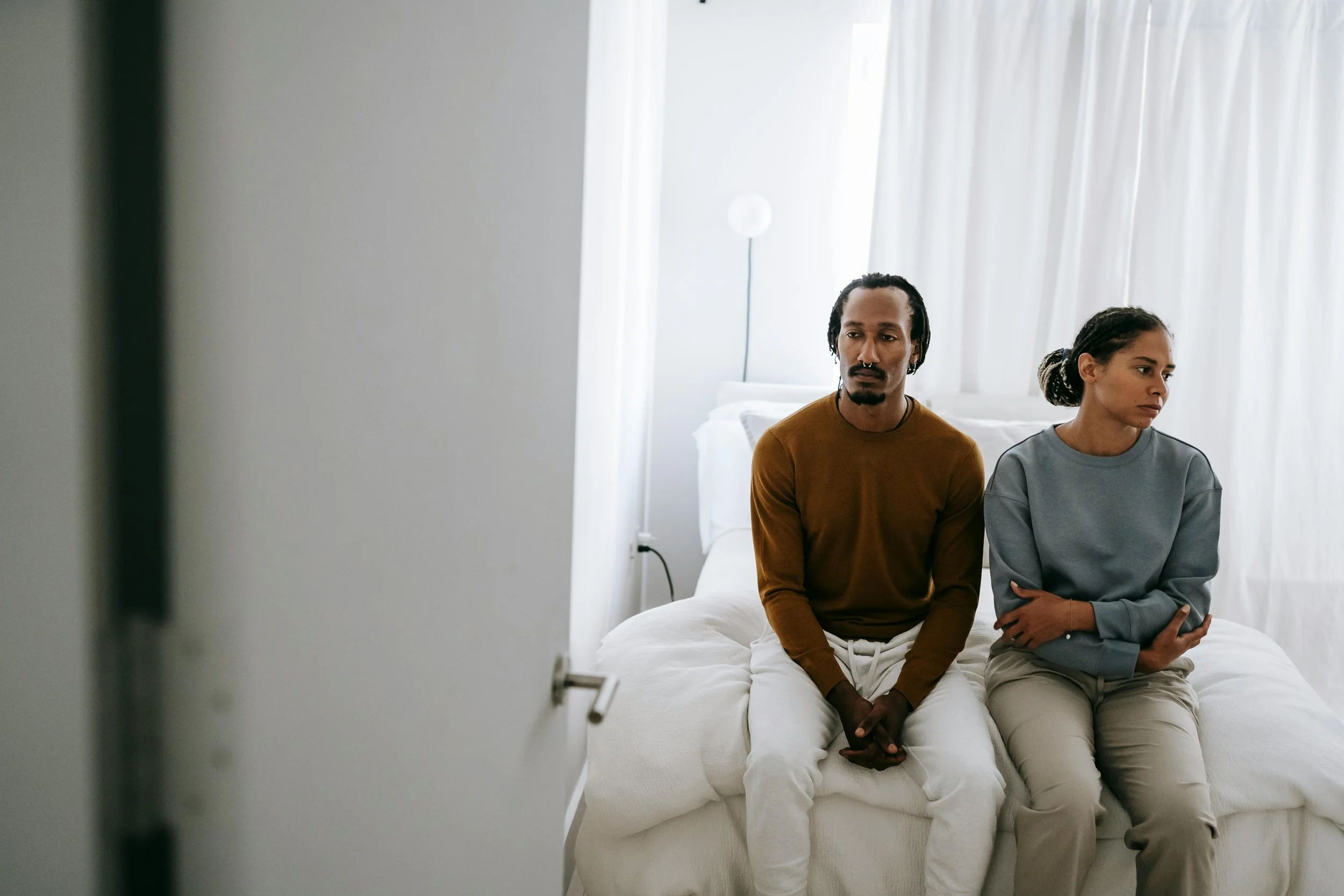 Two people sit on a bed in a bright room with white curtains. A man with dreadlocks and a beard wears a brown shirt, while a woman with braided hair in a gray sweatshirt sits beside him. Both look serious and are looking away from the camera.
