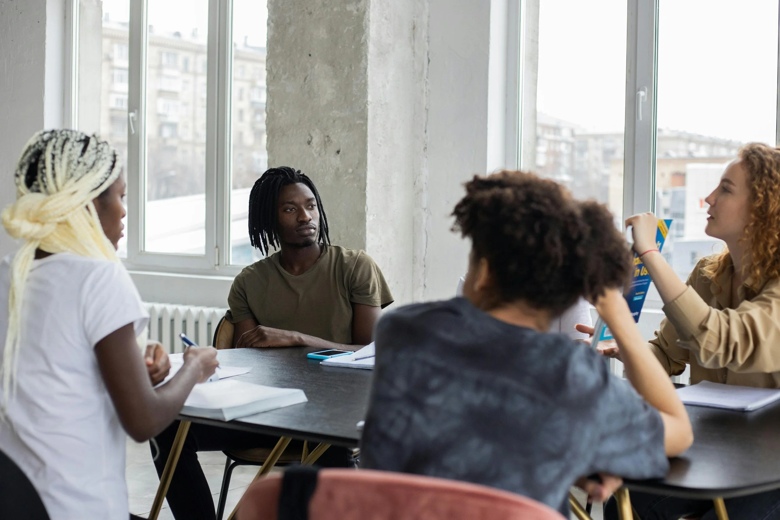Group of diverse young adults having a discussion in a bright, modern room with large windows, some taking notes and one showing a book.