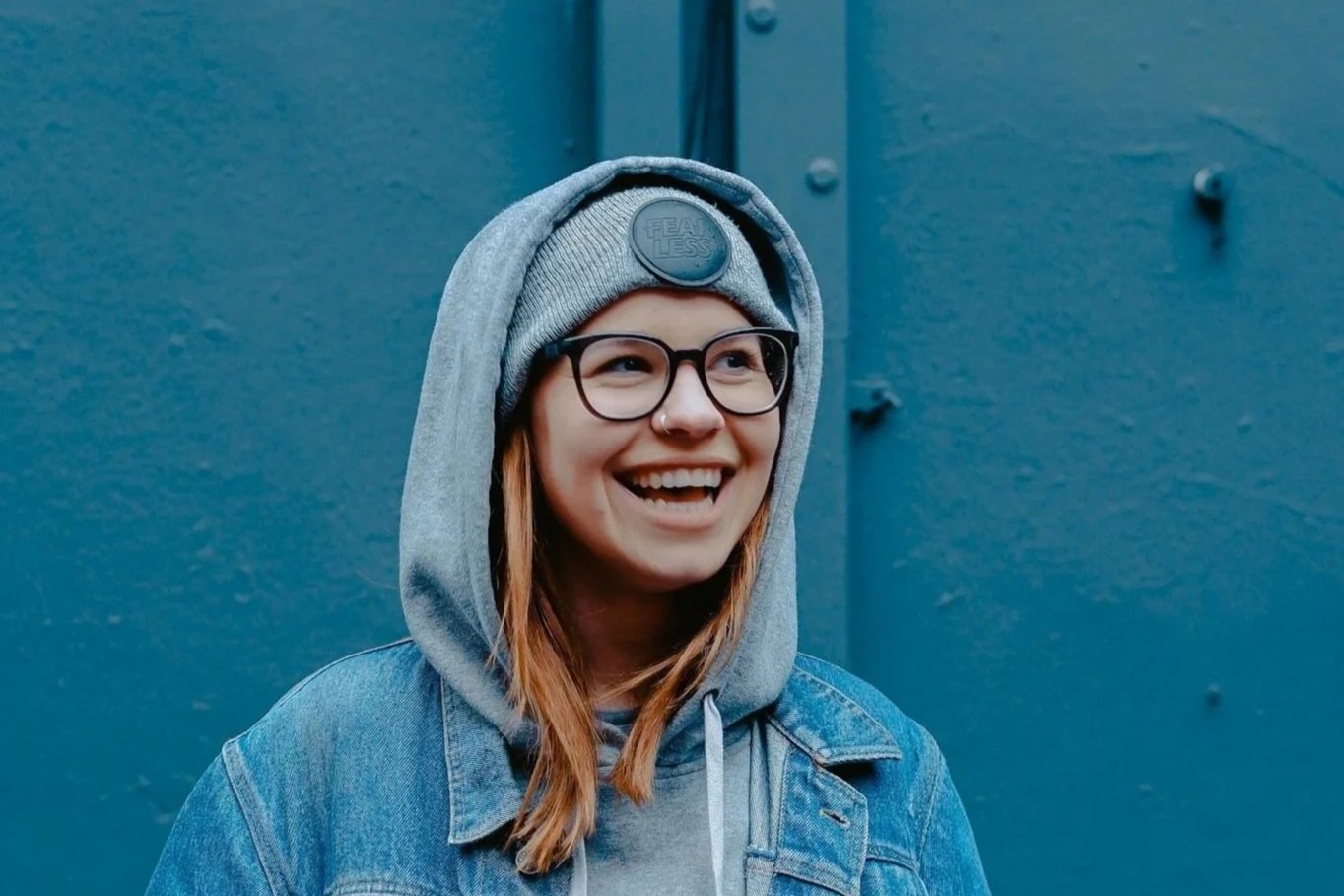 A smiling woman with glasses wearing a grey beanie, hoodie, and denim jacket, standing in front of a blue wall.