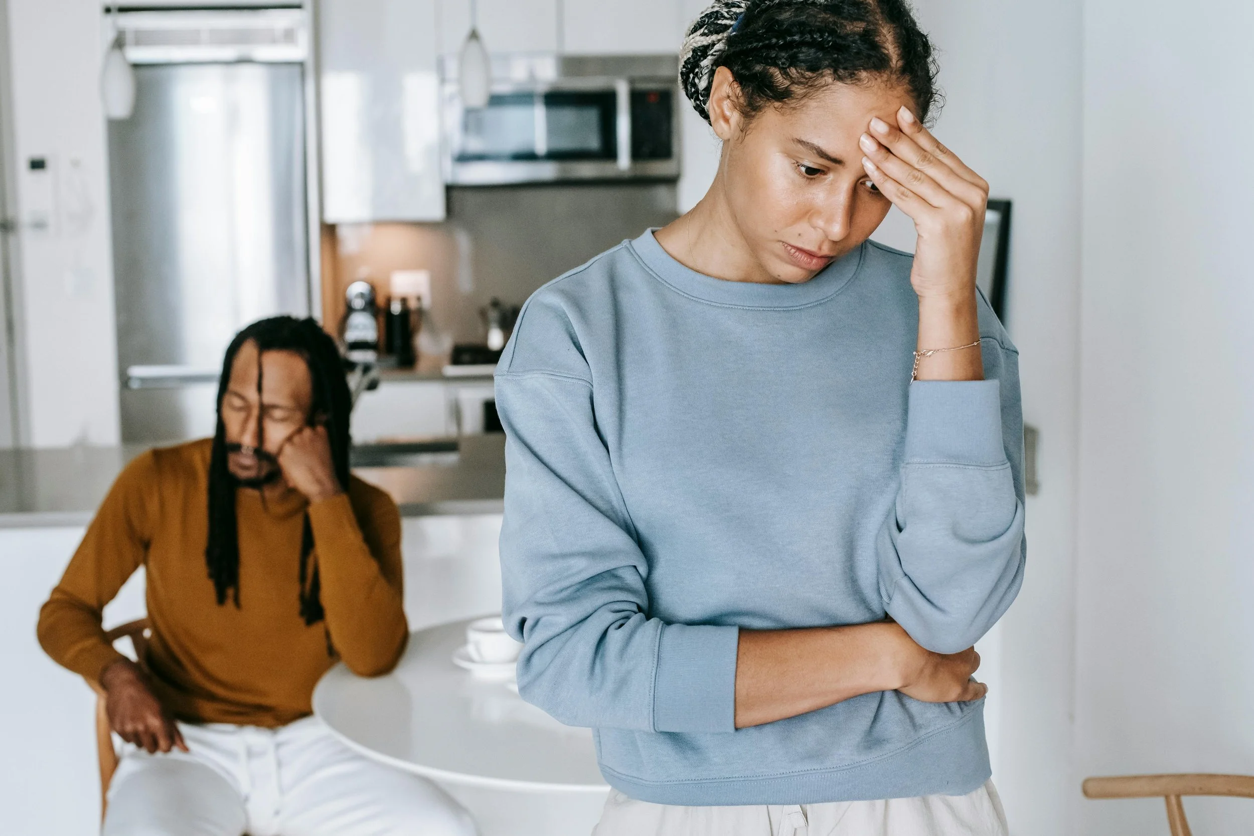 A woman is holding her forehead in a distressed manner while a man in the background appears to be crying or upset.