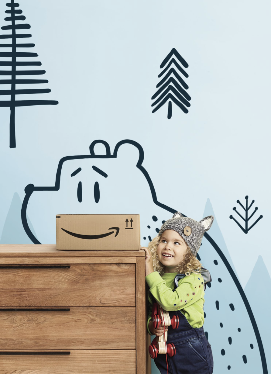 A smiling young girl with curly hair wearing a gray hat with bear ears, a bright green shirt, and dark pants, standing next to a wooden dresser, holding a box with an Amazon logo, in front of a light blue wall decorated with cartoon bear and pine tree illustrations.