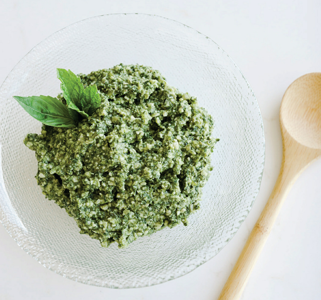 Bowl of green pesto with basil leaves, wooden spoon, and a plain white background.