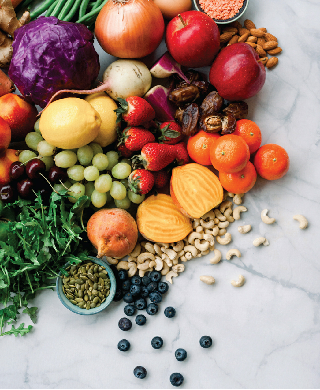 A variety of fresh fruits and vegetables arranged on a marble surface, including grapes, strawberries, apples, oranges, a lemon, a radish, a pomegranate, a head of lettuce, and a small bowl of pumpkin seeds.