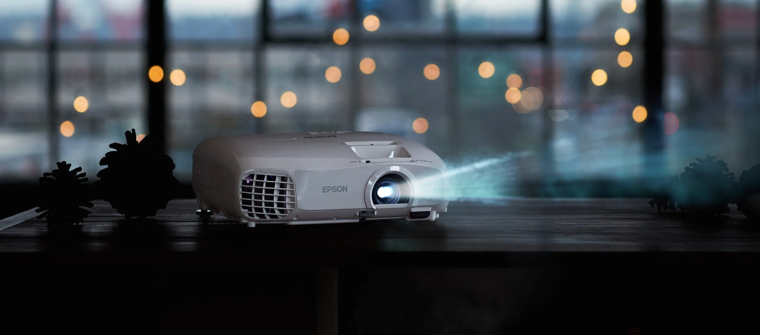 An Epson projector projecting light onto a surface, with pinecone decorations and a blurred window background.