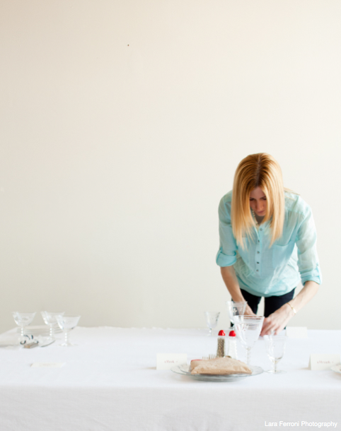 A woman in a light blue shirt setting a table with glassware and a plate in a minimally decorated room.