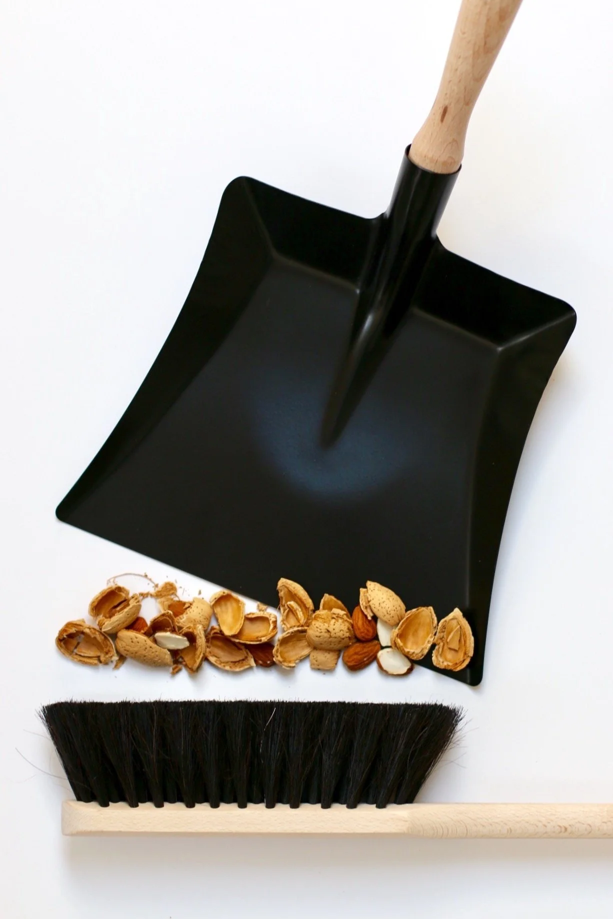 A black dustpan, a broom with black bristles, and a collection of assorted nuts including almonds and walnuts on a white background.
