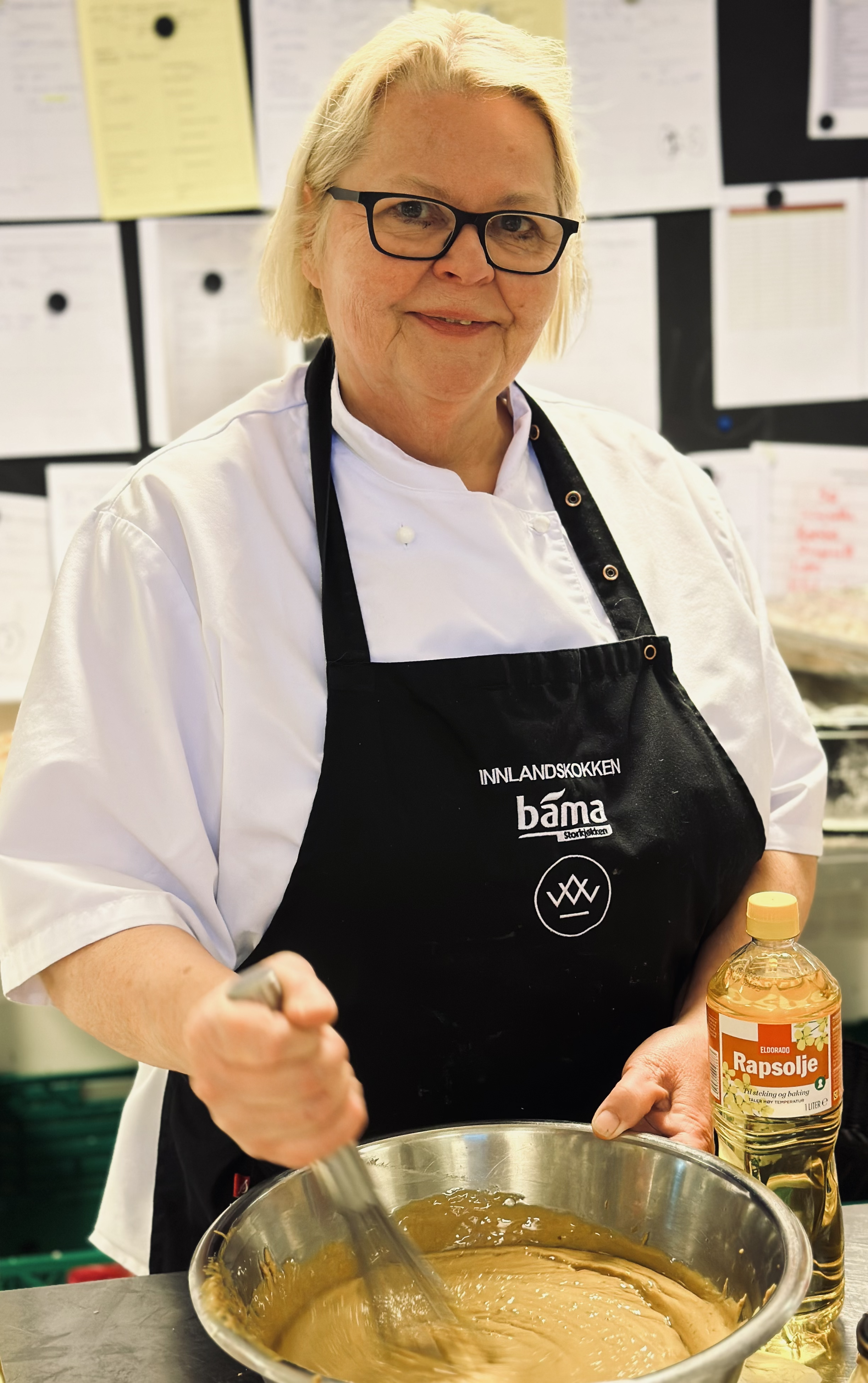En kvinne i kokkekleder rører i en stor bolle med karamellfarge, med en flaske rapsolje ved siden av, i et kjøkken.