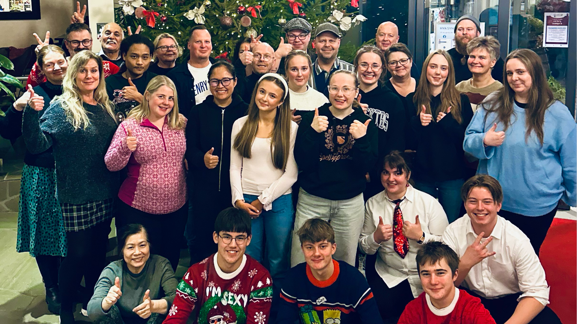En gruppe mennesker foran et juletre, de smiler og gjør julehilsen, noen holder opp tommel opp, andre gjør festlige håndbevegelser.