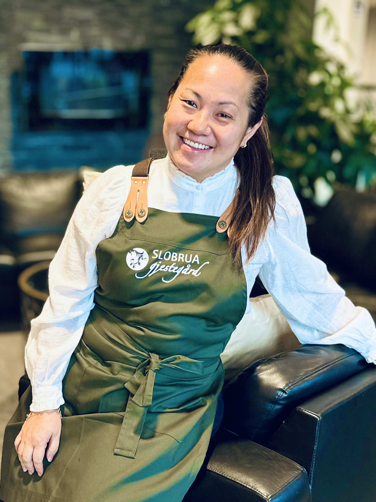 Kvinne smilende i en kafé, iført grønn forkle med teksten 'Slobrua Gjestegård'.