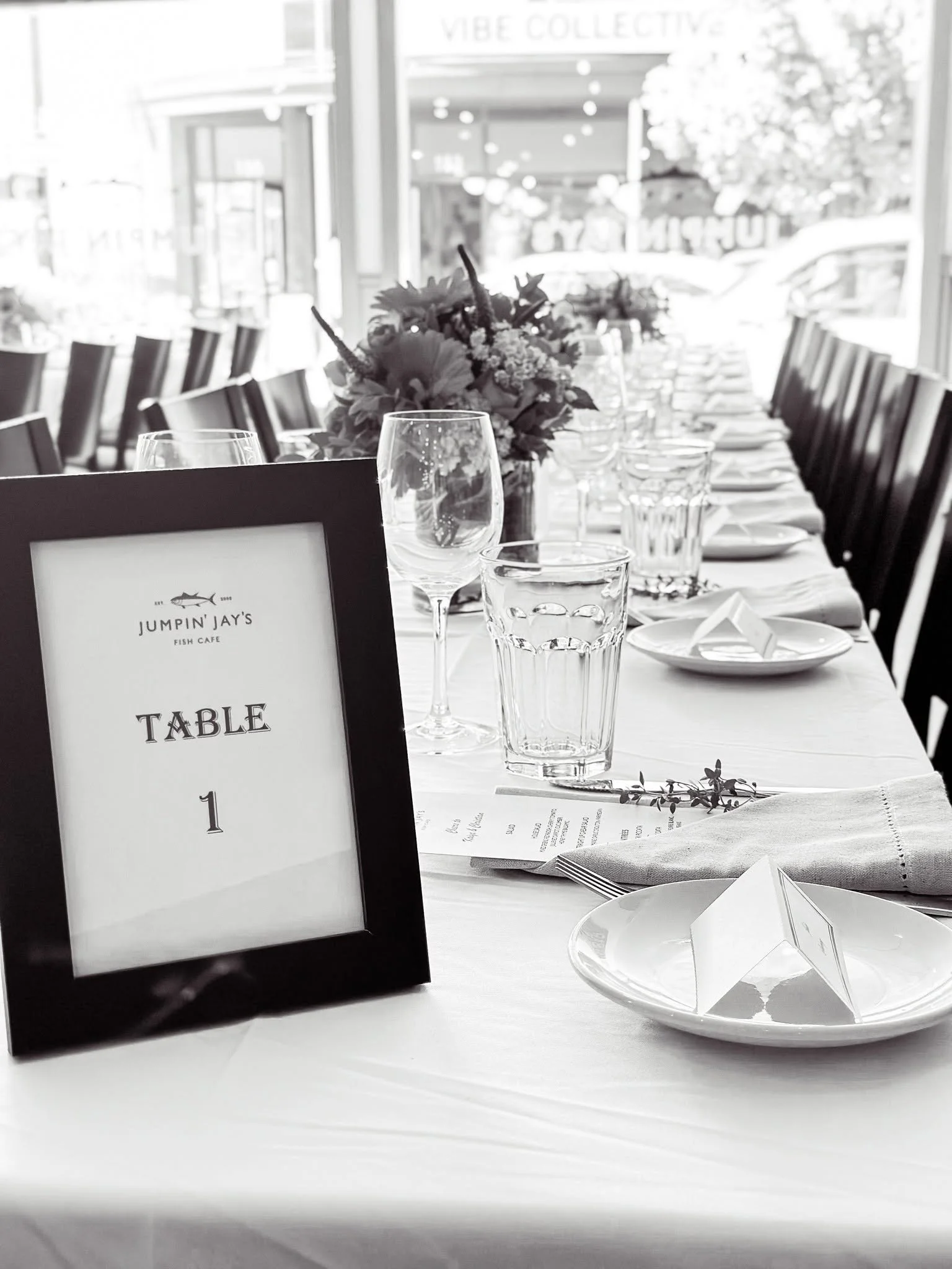 A table set for a meal with a framed menu reading 'Jumpin' Jay's Fish Cafe' and a paper sign indicating 'Table 1.' The table features wine glasses, water glasses, plates, utensils, a floral centerpiece, and place cards, inside a bright restaurant with large windows and a view of trees and parked cars outside.