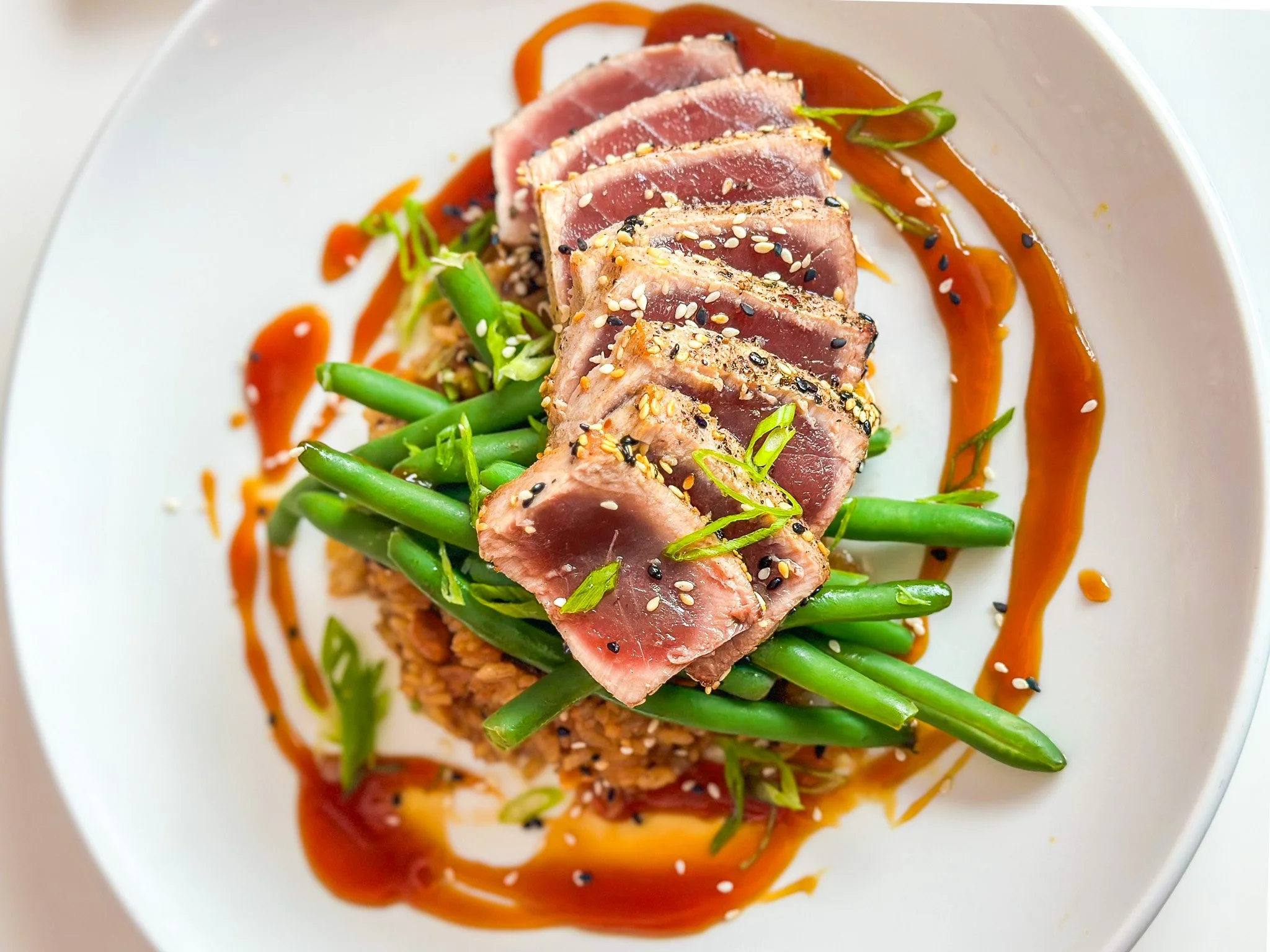 Sliced tuna steak topped with sesame seeds, served on bed of green beans, with a drizzle of sauce and garnished with green onions, on a white plate.