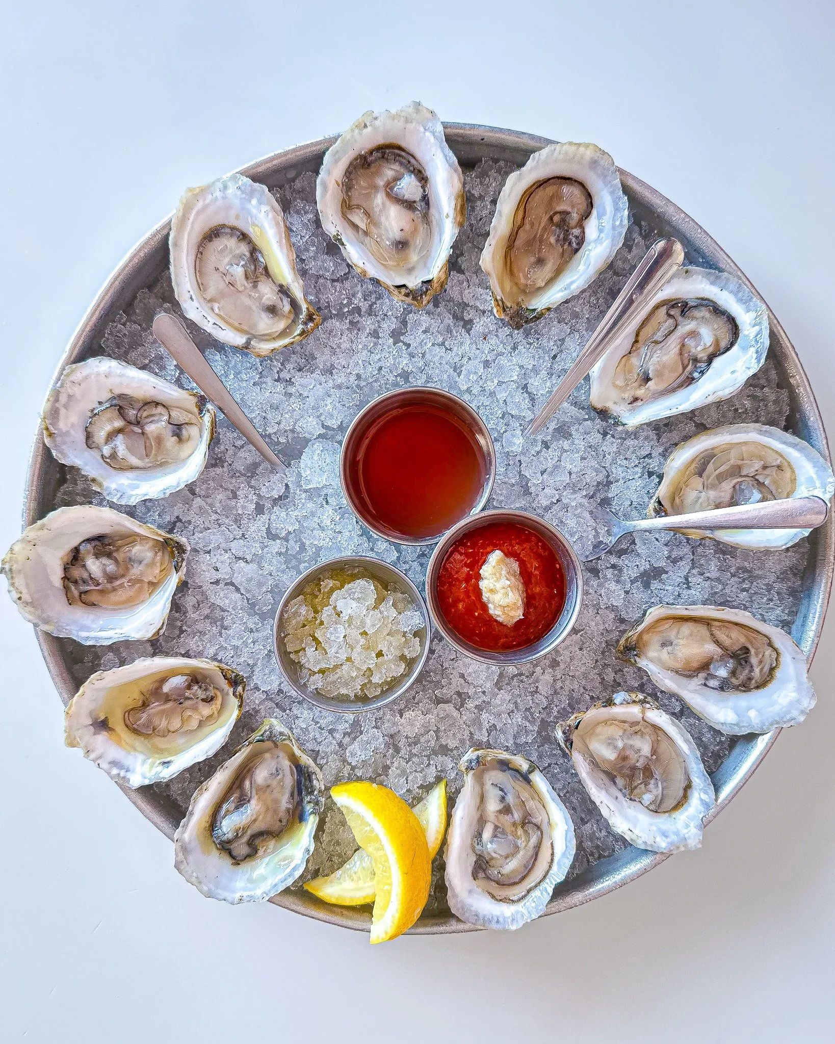 A round metal tray filled with ice and raw oysters on half shells, served with lemon wedges and three small cups of dipping sauces.