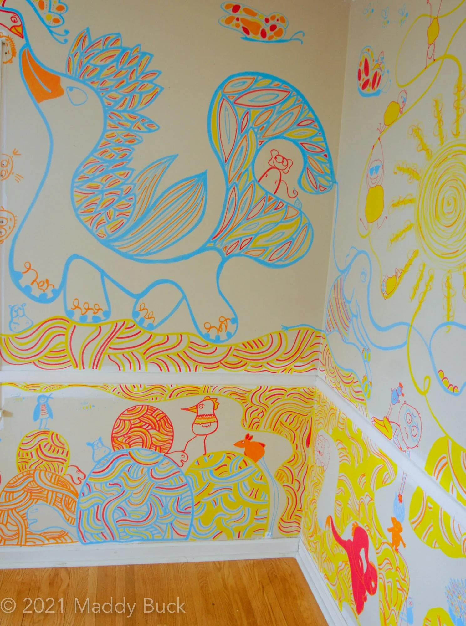  This is a photograph of a mural in a bedroom. We see on top a wild and fantastical dragon creature outlined in blue marker. The creature has lots of features on its back and up its head. It also has a huge windy, wavy tail that sort of has features 