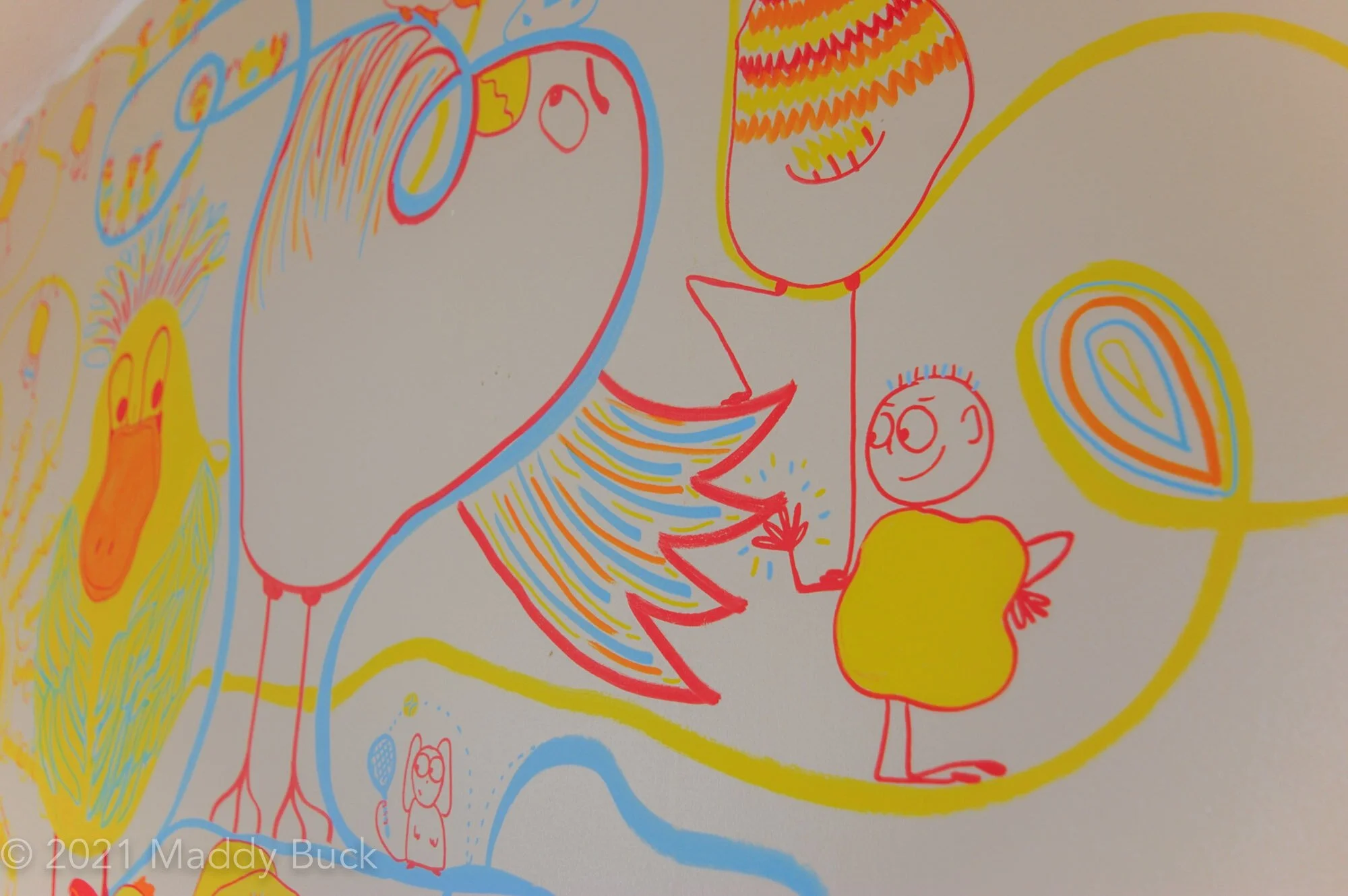  This is a close-up photograph of a mural in a kid bedroom. We see a large bird with its wing outstretch. A blobby kid with a yellow body is on its knees, giving the bird a high five. Standing on top of the bird, we see another bird’s legs and body. 