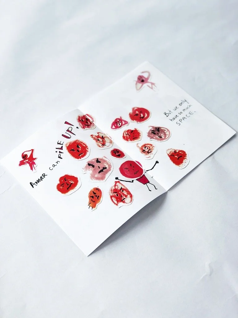  This is a photo of a small picture book, where we can see a bunch of red angry faced blobs. There is text, but we can’t see it.  