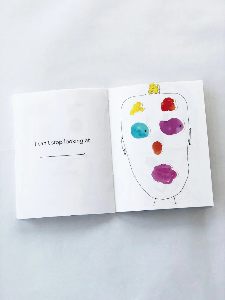  Photo of a picture book open to a page. On the right is a person with a kind of “whatever” face and a buzz cut. The person’s eyes, nose, eyebrows,  mouth, and hair are created with collage pieces, blobs of watercolor paper that have been painted. On