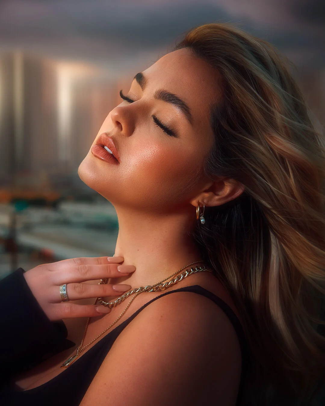 A woman with closed eyes and relaxed expression, wearing jewelry, against a cityscape background.