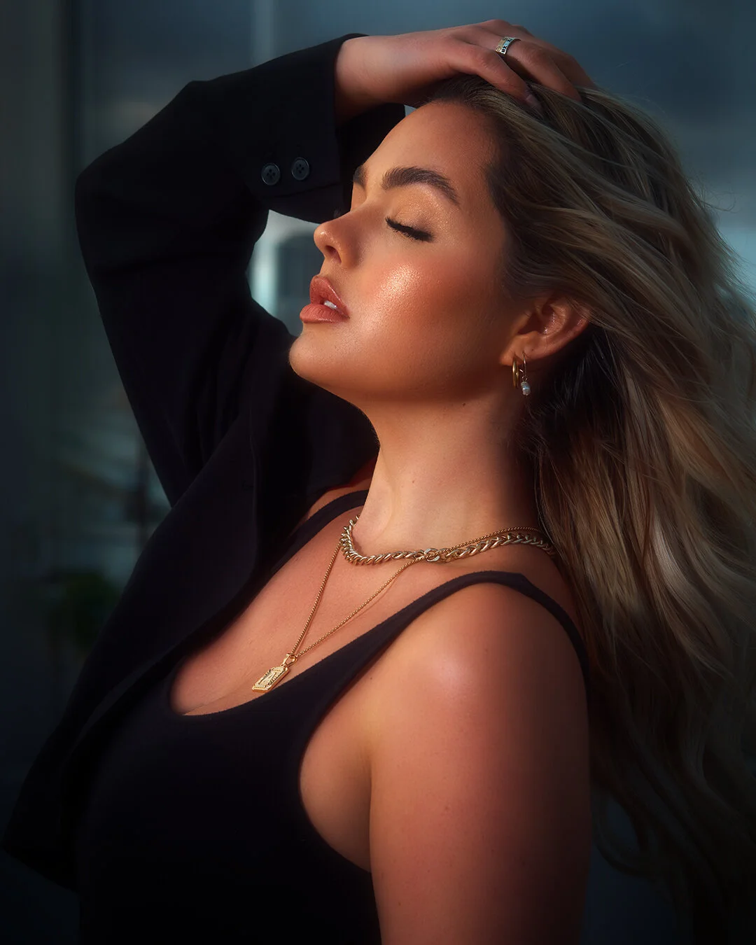 Close-up of a woman with closed eyes, wearing jewelry, with hair flowing, in a black tank top and jacket, with a blurred cityscape background.