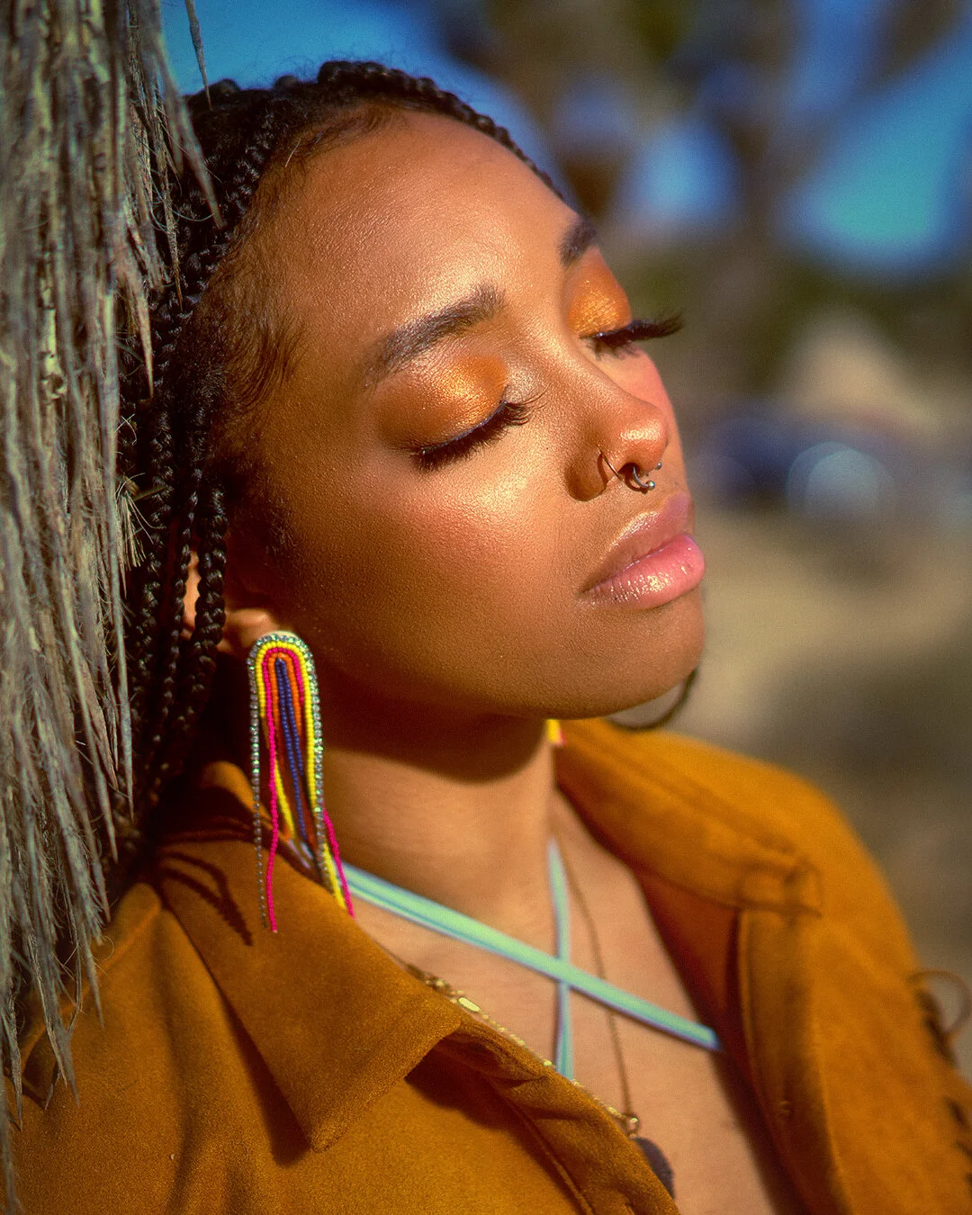 Close-up of a young woman with dark braided hair and gold eye makeup, wearing colorful beaded earrings and a mustard-colored jacket, with her eyes closed and leaning against a tree