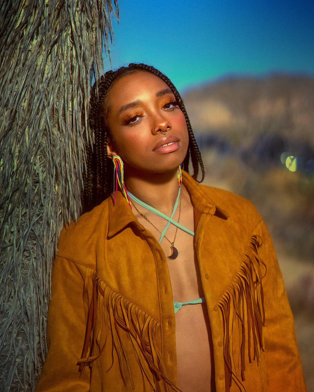 A woman with braided hair, wearing a brown fringed suede jacket, colorful earrings, and a moon necklace, leaning against a palm tree at sunset or dusk.