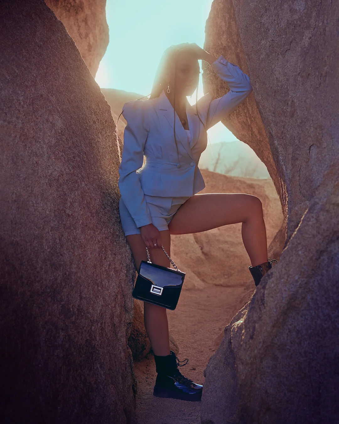 Woman in a beige suit with shorts, black boots, and a black handbag posing among large rock formations with sunlight behind her.