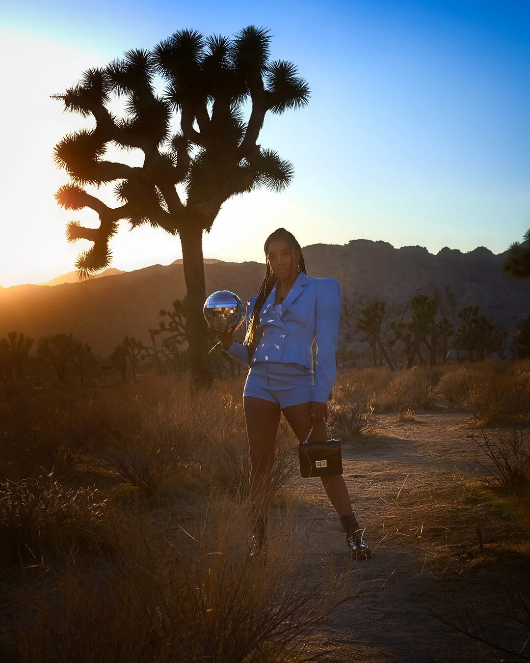 A woman in a light blue suit holding a disco ball in a desert landscape at sunset, with Joshua trees in the background.