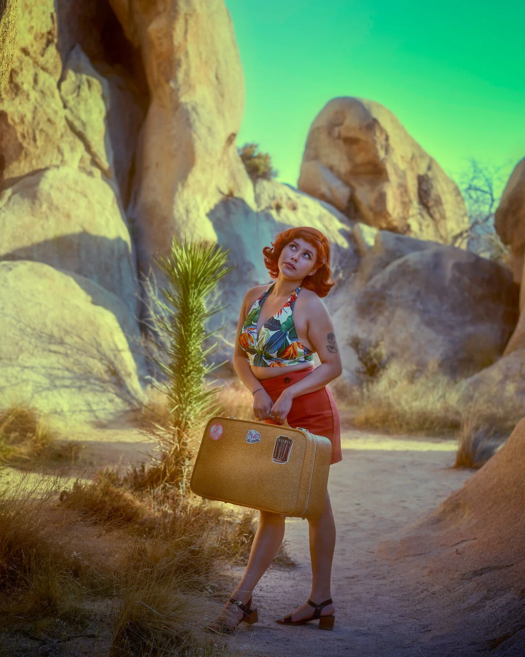 A woman with red hair wearing a tropical print top and red skirt, carrying a suitcase, standing in a rocky desert landscape with large boulders and sparse vegetation.