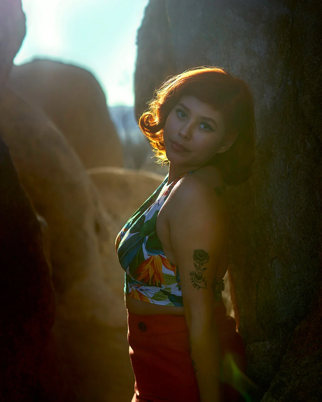 A woman with short red hair, wearing a colorful floral top and red pants, leaning against a rock with sunlight illuminating her face and hair, and a tattoo visible on her left arm.