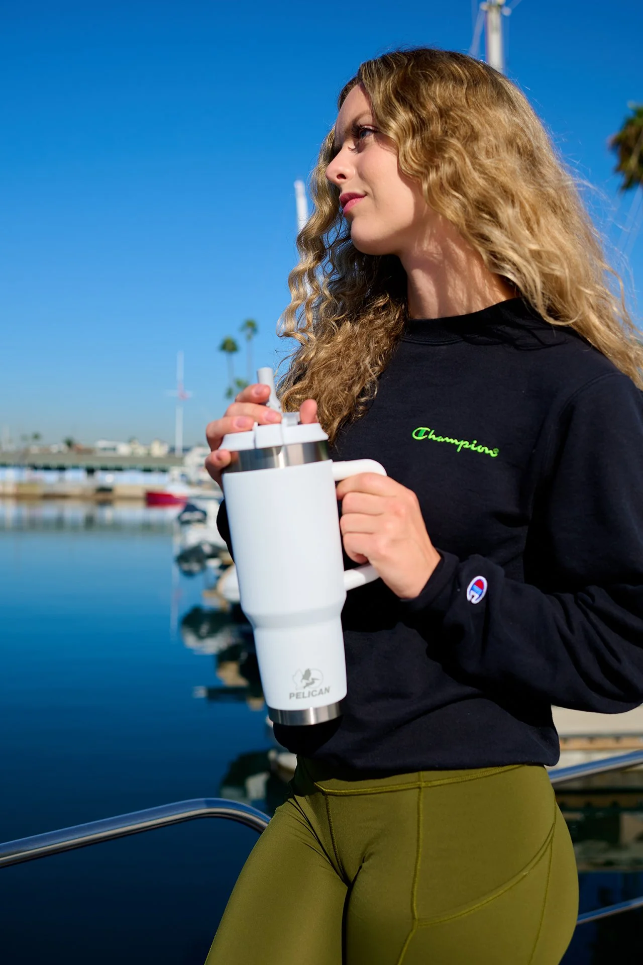 A woman with curly blonde hair holding a white portable blender near a marina with boats and water in the background, wearing a black sweatshirt with the word 'Champion' embroidered on it and olive green pants.