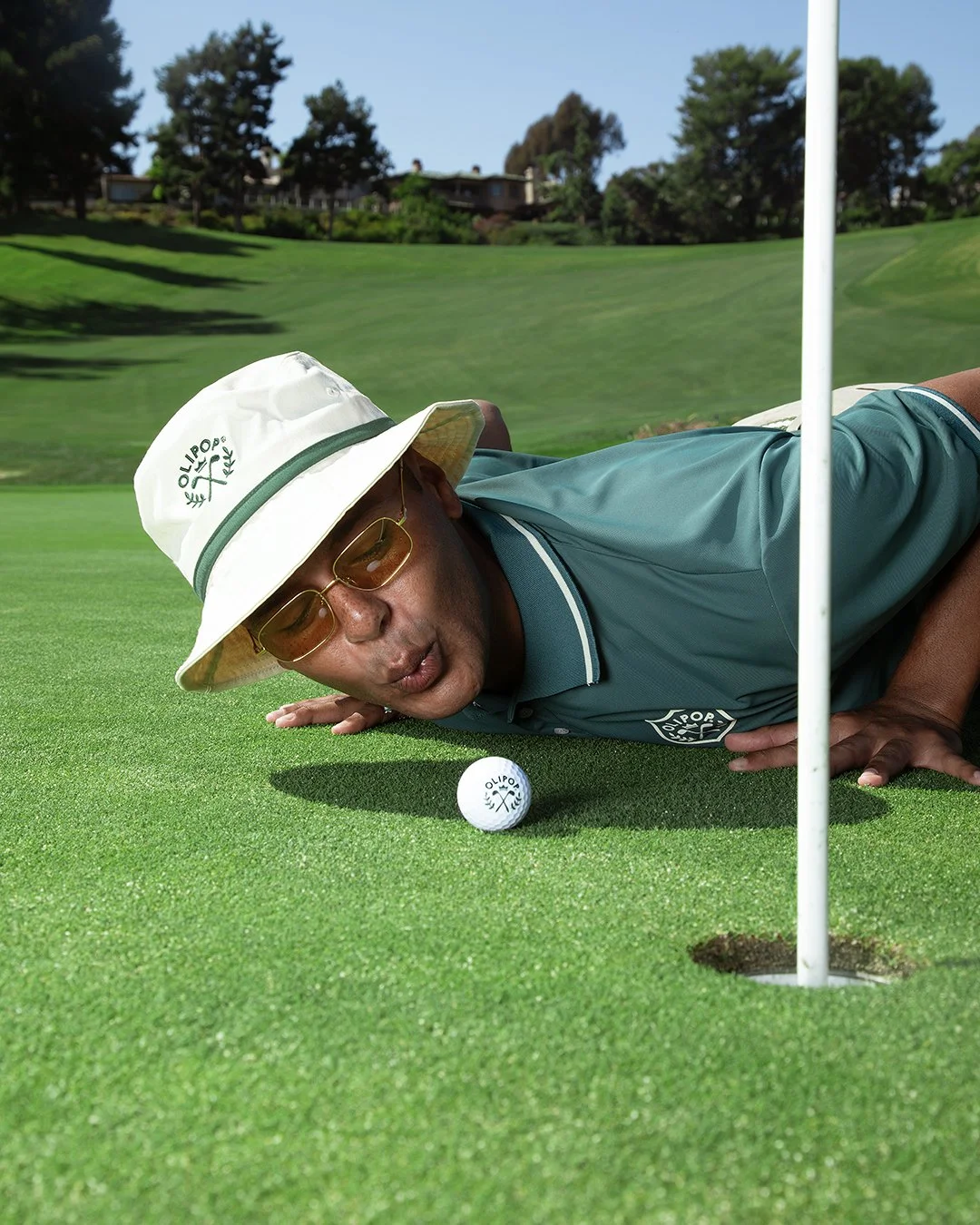 A man lying on a golf green near a hole, wearing a white bucket hat with 'OLIPOP' logo, sunglasses, and a teal golf shirt, about to hit a golf ball.