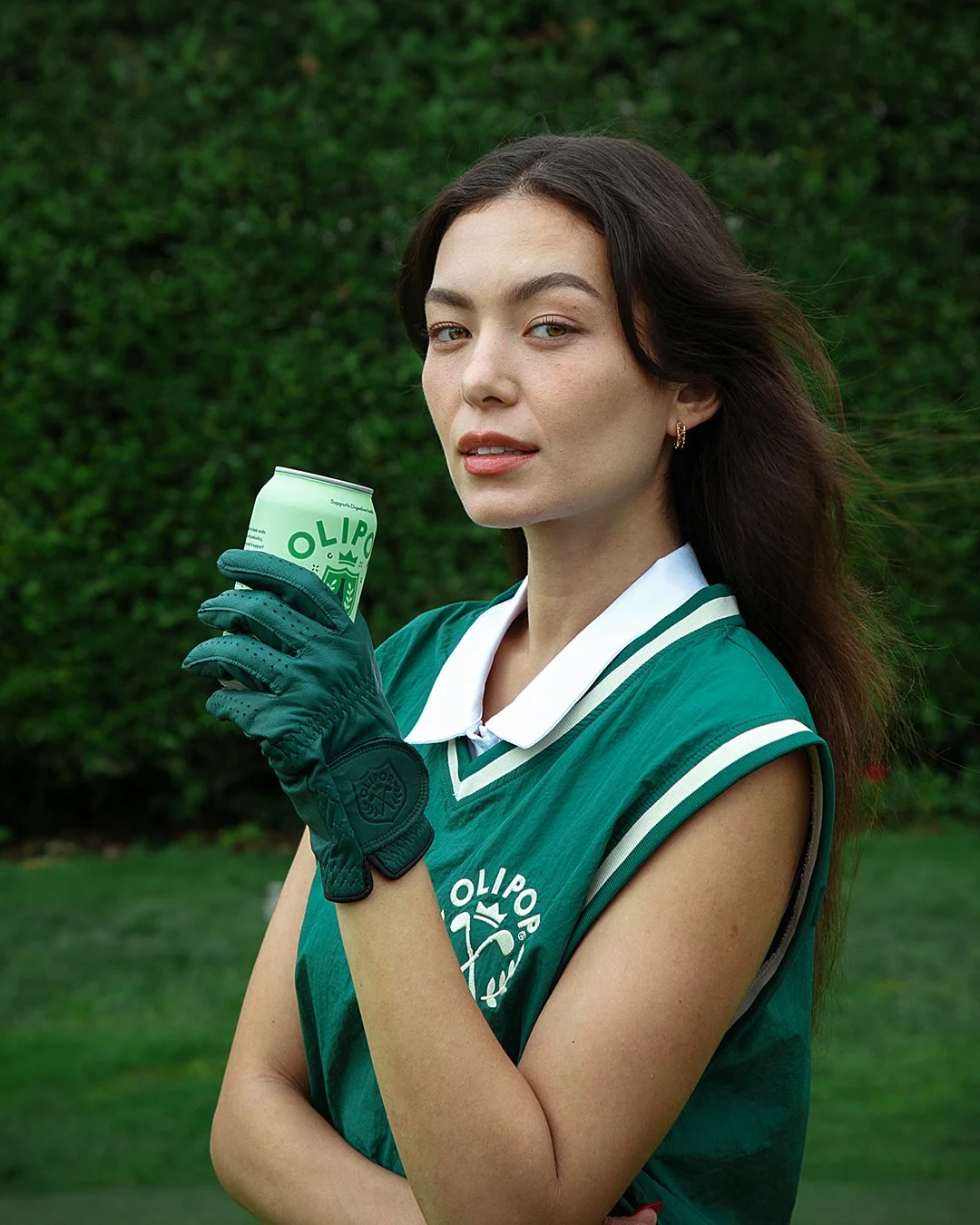 A young woman with long dark hair and fair skin holding a green can, wearing a green sports vest with white trim and a white collar, with green gloves, standing outdoors in front of a hedge.
